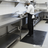 Person washing dishes in a commercial kitchen with a black scraper mat on the floor.