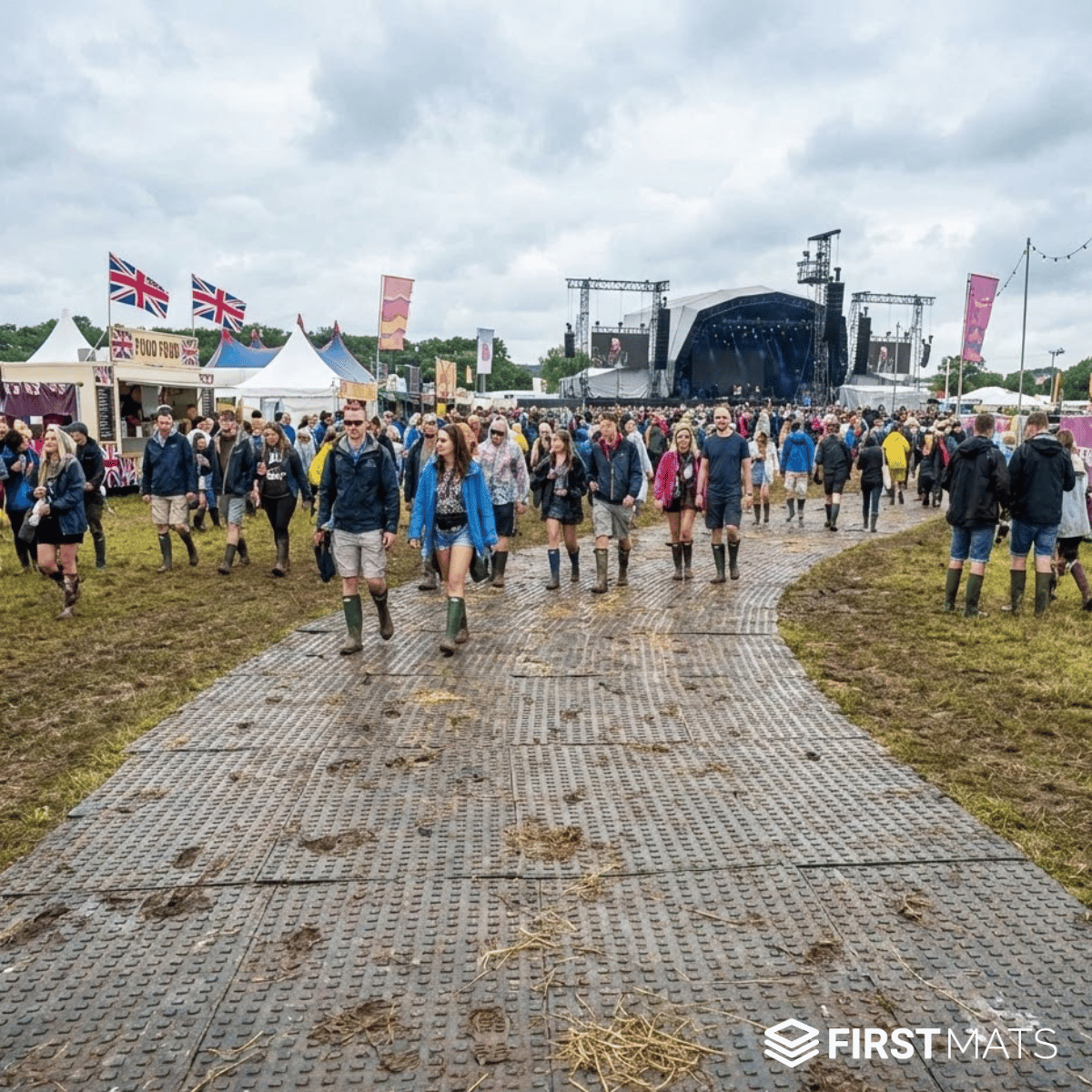 People walking on ground protection mats at a festival with tents and stage in the background
