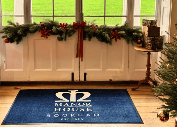 Blue logo mat with 'Manor House Bookham' logo in a festive room with Christmas decorations.