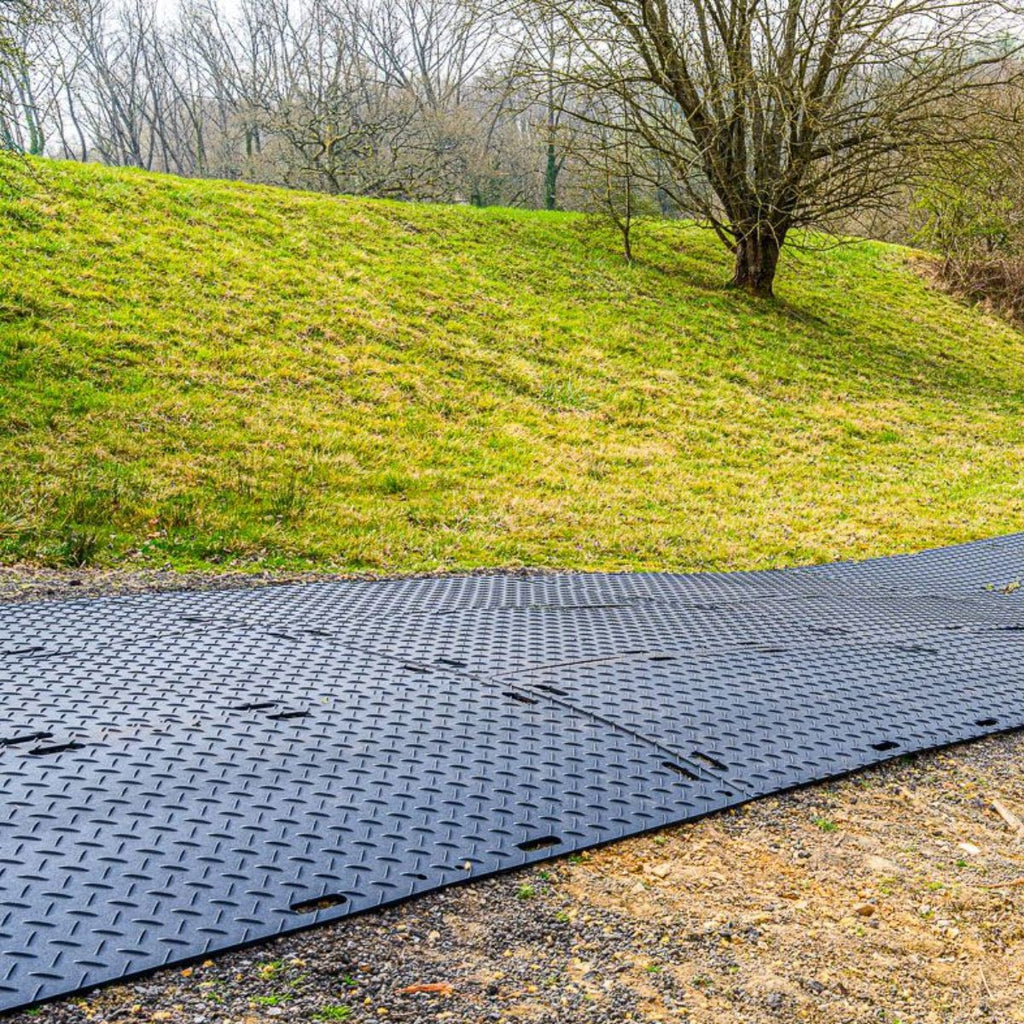 Black temporary car park mats on a gravel surface with a grassy hill and tree in the background