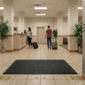 People with luggage walking through a lobby with a black mat on the floor.