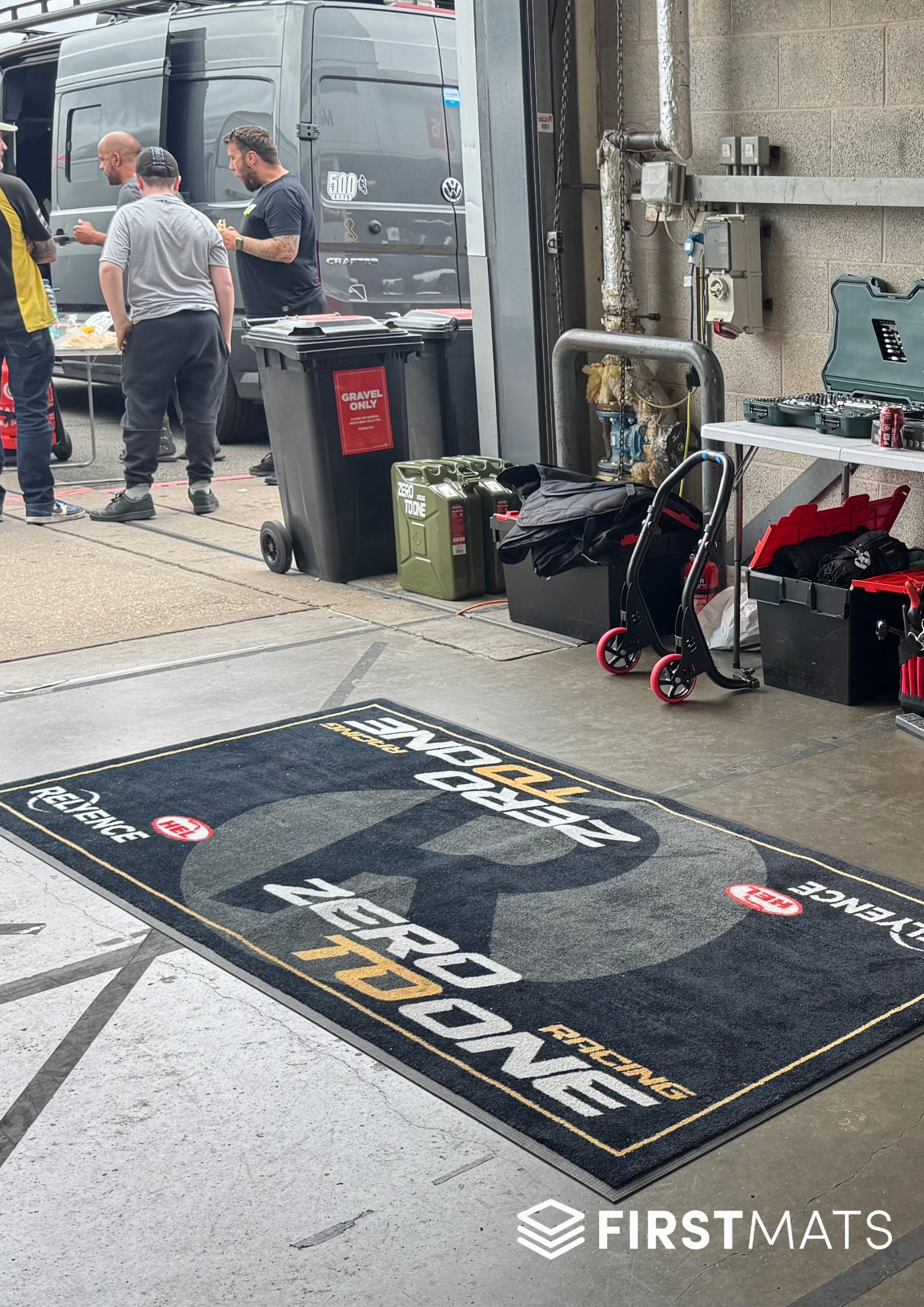Promotional mat with branding on a concrete floor, featuring 'FirstMats' and 'Zero to One Racing'.